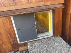 En grå Omlet automatisk døråbner til hønsegård, der er fastgjort til en hønsegård i træ.
