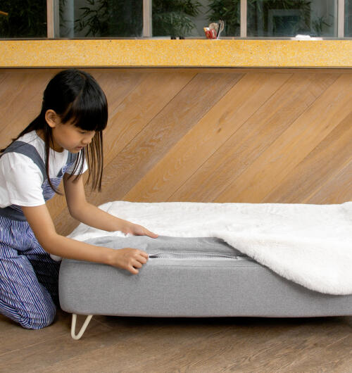 Girl Zipping a white faux sheepskin topper onto Topology dog bed madress with white hairpin feet.